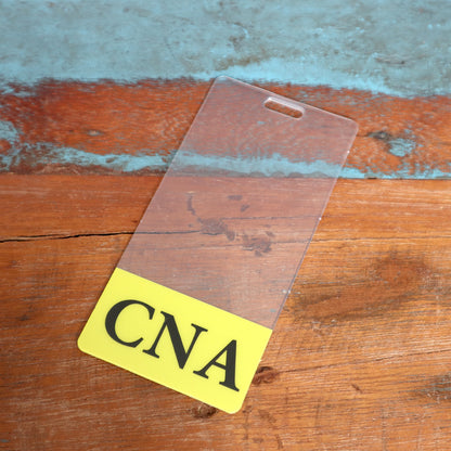 A transparent ID badge with "CNA" printed in bold black letters on a yellow strip lies on a wooden surface, complemented by a Clear CNA Badge Buddy Vertical with Yellow Border for Nursing Assistants to assist nursing assistants.