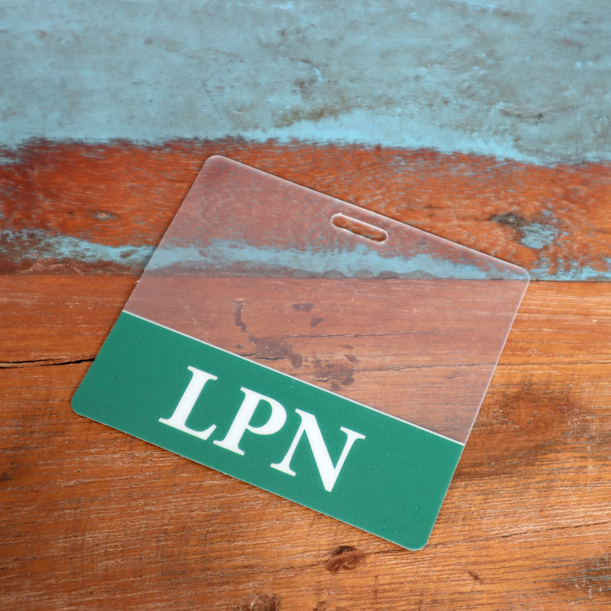 Close-up of a plastic badge lying on a wooden surface. The Clear LPN Badge Buddy - Horizontal ID Badge Backer for Licensed Practical Nurses - Double Sided Print features a green section at the bottom with white letters reading "LPN", perfect for licensed practical nurses in any healthcare setting.