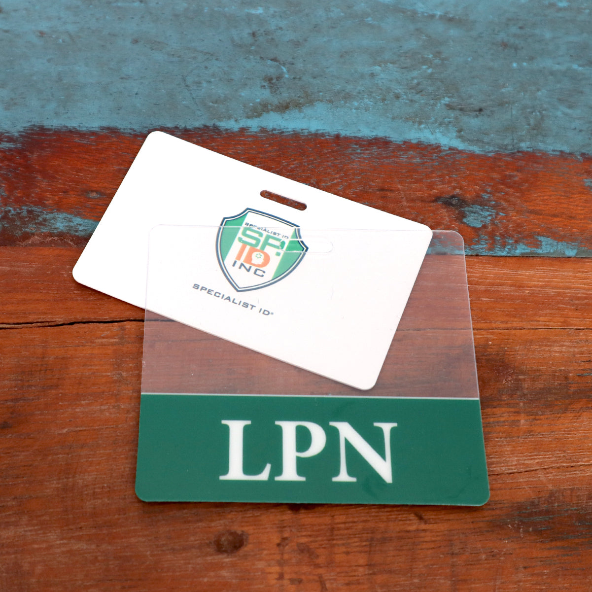 Two identification cards are placed against a weathered wooden surface. One card has "LPN" written at the bottom, clearly displaying a Clear LPN Badge Buddy - Horizontal ID Badge Backer for Licensed Practical Nurses - Double Sided Print, while the other card showcases a logo and the text "Specialist ID." This setup is ideal for any healthcare setting.