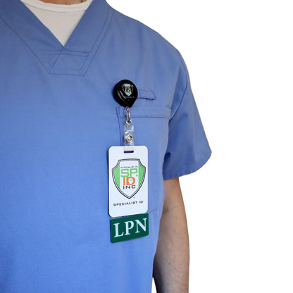 A person is wearing a blue medical scrub top with a Clear LPN Badge Buddy Vertical with Green Border for Licensed Practical Nurses clipped to the chest pocket that reads "LPN. Sold by Specialist ID.