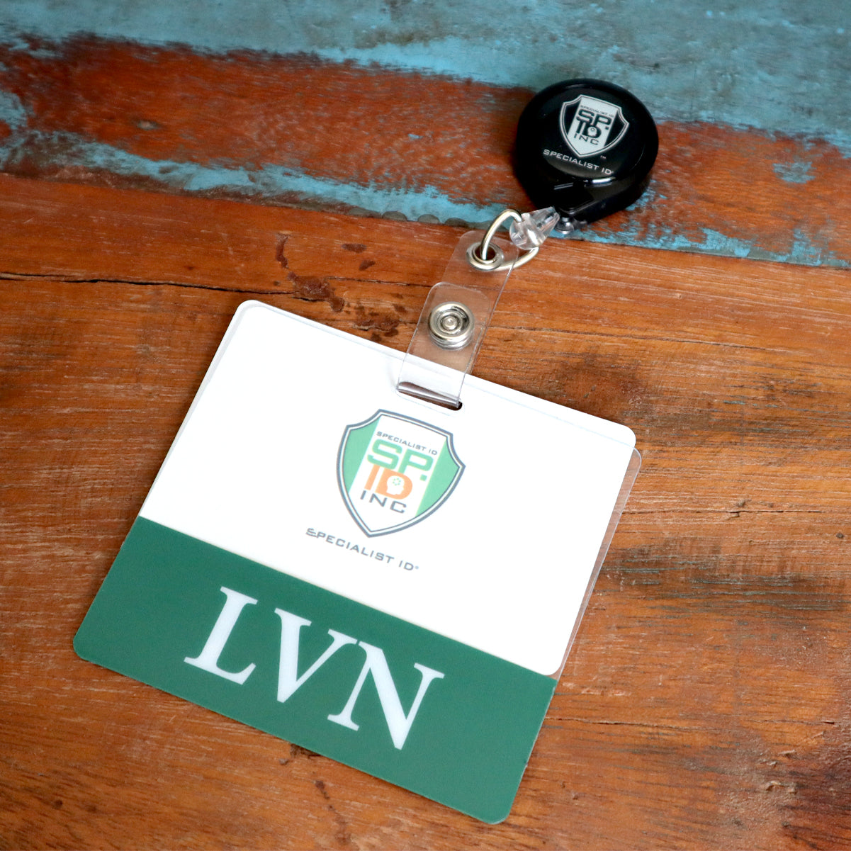 A badge with the letters "LVN," a security emblem, and a retractable nurse badge holder lying on a wooden surface, perfectly complemented by a Clear LVN Badge Buddy - Horizontal ID Badge Backer for Licensed Vocational Nurses - Double Sided Print.