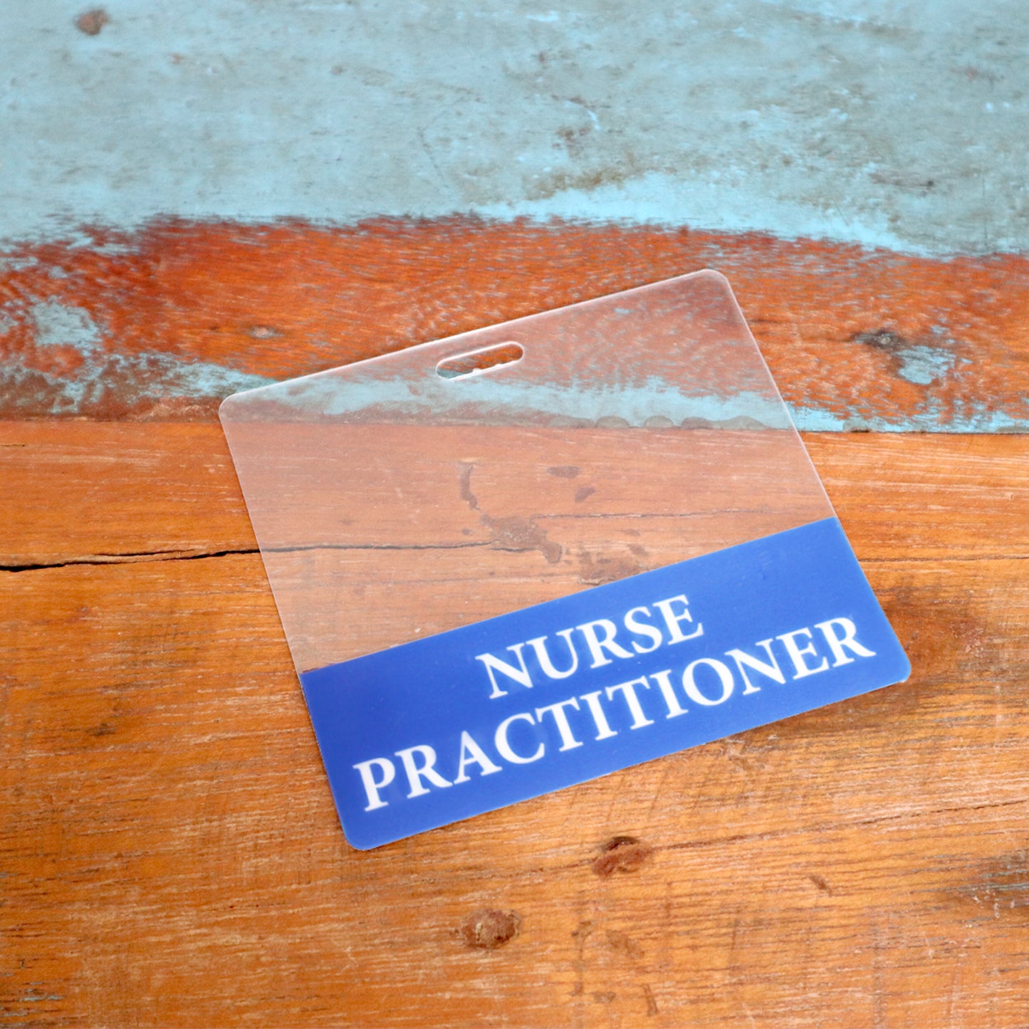 The Clear NURSE PRACTITIONER Badge Buddy, a horizontal double-sided ID badge backer, features "Nurse Practitioner" text on a blue and orange background and is shown resting on a wooden surface.