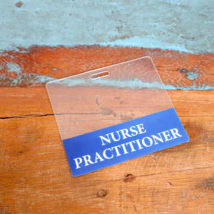 The Clear NURSE PRACTITIONER Badge Buddy, a horizontal double-sided ID badge backer, features "Nurse Practitioner" text on a blue and orange background and is shown resting on a wooden surface.