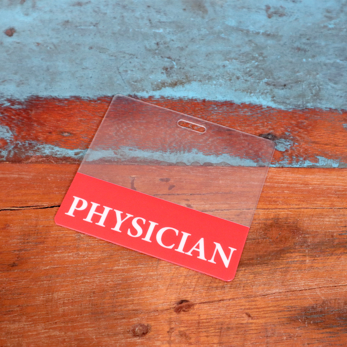 A Clear PHYSICIAN Badge Buddy - Horizontal ID Badge Backer for Physicians - Double Sided Print with a red color border lies on a wooden surface with a blue background.