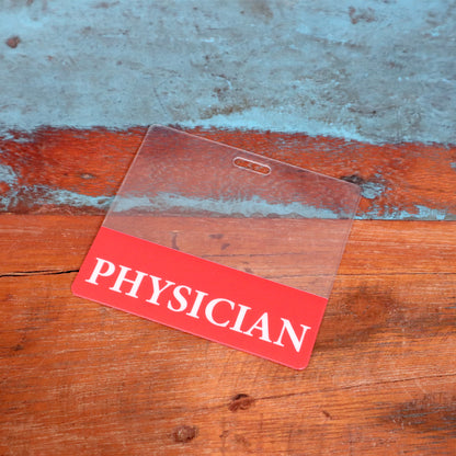 A Clear PHYSICIAN Badge Buddy - Horizontal ID Badge Backer for Physicians - Double Sided Print with a red color border lies on a wooden surface with a blue background. Sold by Specialist ID.