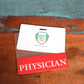 A Clear PHYSICIAN Badge Buddy - Horizontal ID Badge Backer for Physicians - Double Sided Print is placed on a wooden surface with a blue and brown background.