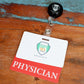 A plastic badge with a retractable holder labeled "PHYSICIAN" is displayed on a wooden surface. The Clear PHYSICIAN Badge Buddy - Horizontal ID Badge Backer for Physicians - Double Sided Print features the logo of Specialist ID with green, yellow, and white colors.