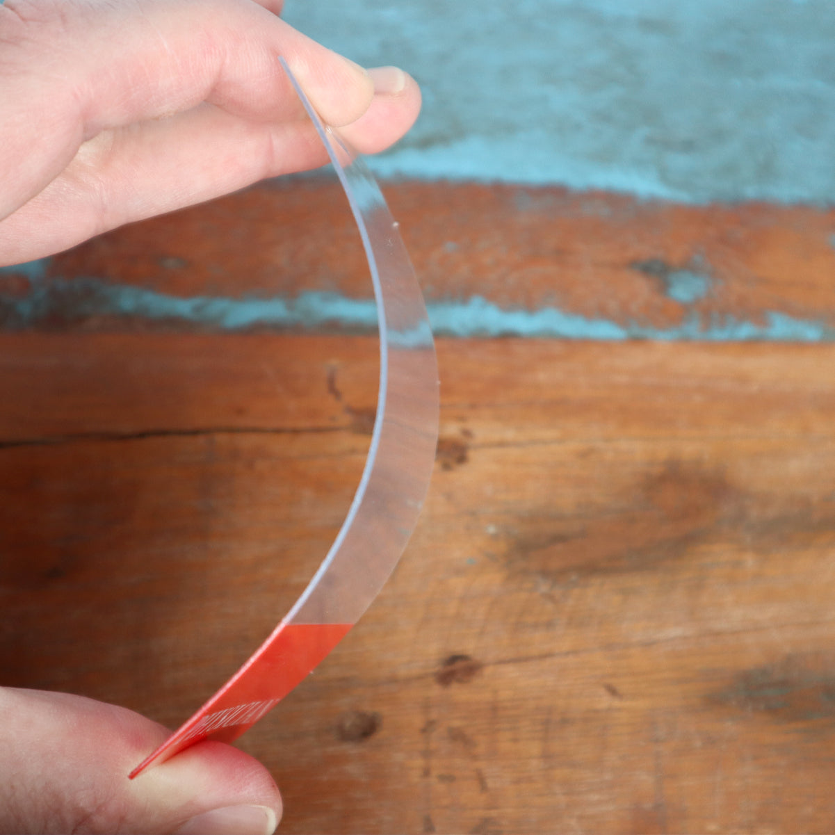 A person holds a thin, flexible piece of clear plastic with a red base, similar to Custom Printed Clear Badge Buddies Vertical (Standard Size) - Customize with Title and Color, showing its bendability over a wooden surface.