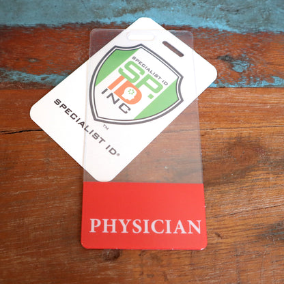 A clear badge holder contains a card with a shield logo and the text "SPECIALIST ID" and "SP INC." Below is a **Clear PHYSICIAN Badge Buddy Vertical with Red Border for Physicians**. Both items lie on a wooden surface.