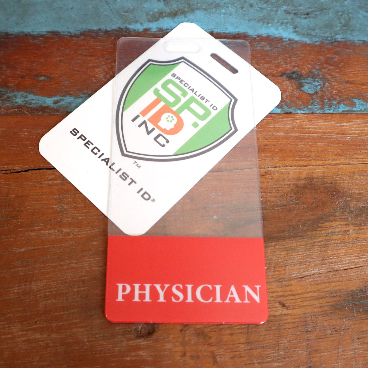 A red and white badge labeled "PHYSICIAN" lies on a wooden surface, with a Specialist ID card partially covering it. These Custom Printed Clear Badge Buddies Vertical (Standard Size) - Customize with Title and Color provide instant role recognition in any medical setting.