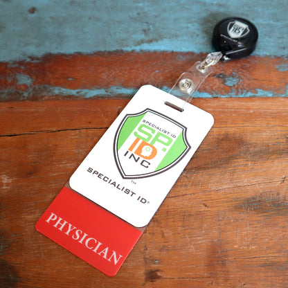 A Clear PHYSICIAN Badge Buddy Vertical with Red Border for Physicians with a Specialist ID Inc lanyard reel, placed on a wooden surface, snugly fits in a clear badge holder. The badge has a red section labeled "PHYSICIAN" and a white section with a green and orange logo.