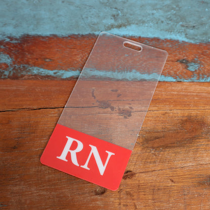 A Clear RN Badge Buddy Vertical with Color Border for Registered Nurses - Double Sided Print with a red bottom section displaying the letters "RN" in white lies on a rustic wooden surface with peeling blue paint, perfect for nurses seeking a vertical ID backer.