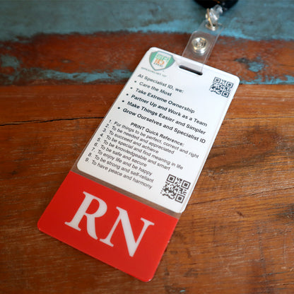 A white and red vertical ID backer with "RN" at the bottom, listing principles including ownership, teamwork, simplification, and boosting morale. This Clear RN Badge Buddy Vertical with Color Border for Registered Nurses - Double Sided Print is attached to a lanyard clip on a wooden surface, perfect for nurses committed to excellence.