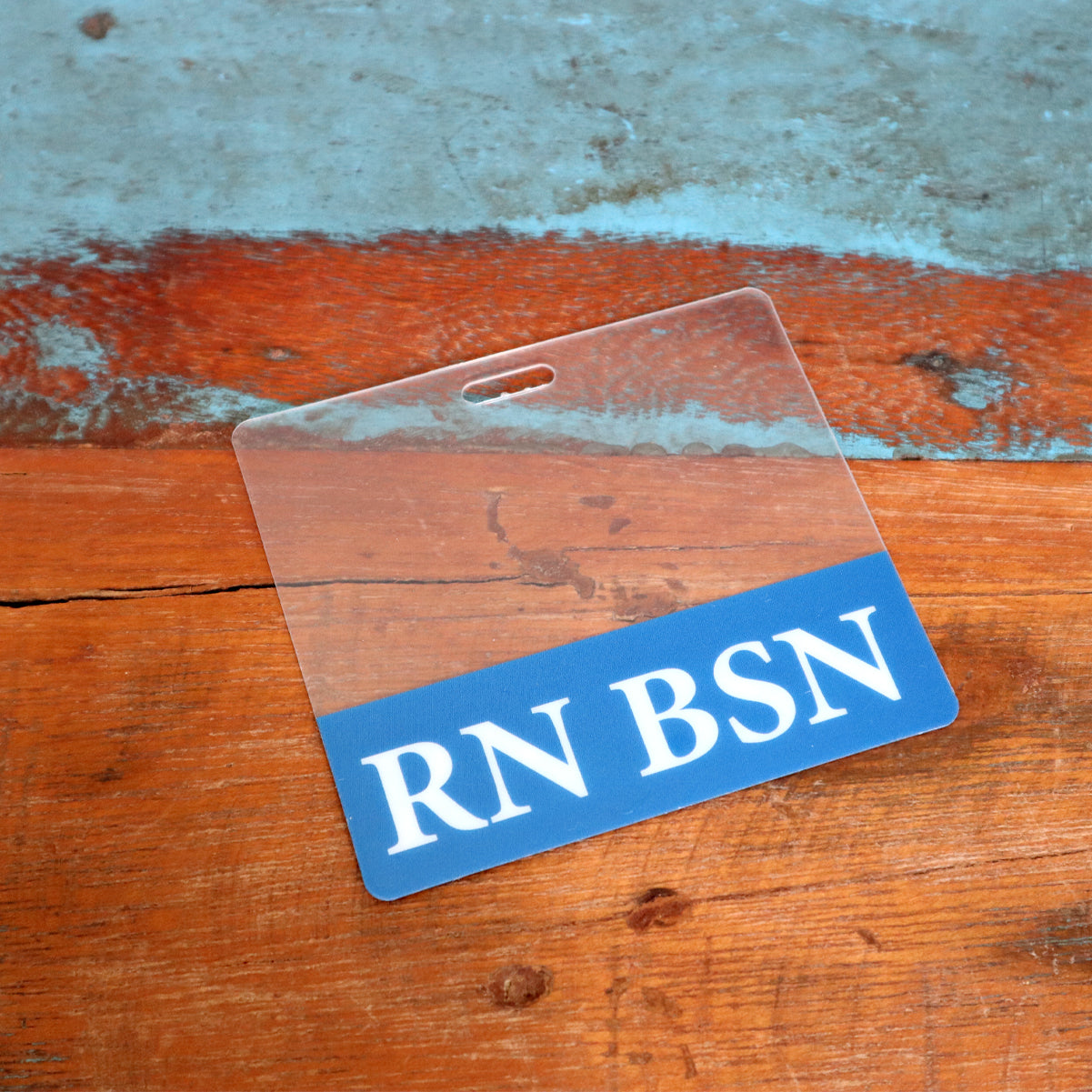 A Clear RN BSN Badge Buddy - Horizontal Nurse ID Badge Backer - Double Sided Print is placed on a wooden surface with a weathered, painted area above.