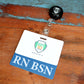 A name badge with "RN BSN" at the bottom and an attached retractable badge holder, placed on a wooden surface. Perfect for nurses with a Bachelor of Science in Nursing, this Clear RN BSN Badge Buddy - Horizontal Nurse ID Badge Backer - Double Sided Print adds a professional touch to your uniform.
