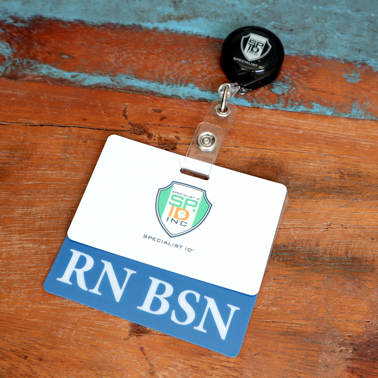 A name badge with "RN BSN" at the bottom and an attached retractable badge holder, placed on a wooden surface. Perfect for nurses with a Bachelor of Science in Nursing, this Clear RN BSN Badge Buddy - Horizontal Nurse ID Badge Backer - Double Sided Print adds a professional touch to your uniform.