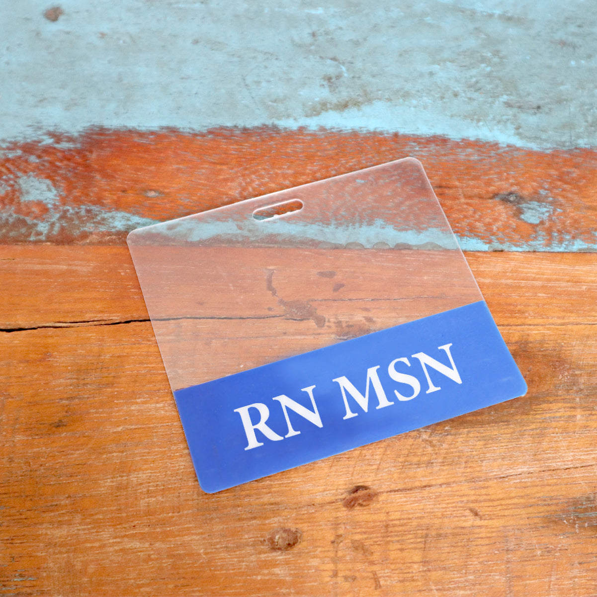A Clear RN MSN Badge Buddy - Horizontal Nurse ID Badge Backer with double-sided print is shown on a wooden surface painted in blue and orange hues. Sold by Specialist ID.