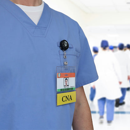 Close-up of a person wearing blue scrubs and a CNA badge with a photo ID attached, while healthcare professionals walk in the background. The badge is secured in one of the CNA BadgeBottom Badge Holder & CNA Badge Buddy IN ONE!! - Horizontal ID Badge Sleeve with Bottom Role Tag for Nurse Assistants, ensuring visibility and security during their busy day.