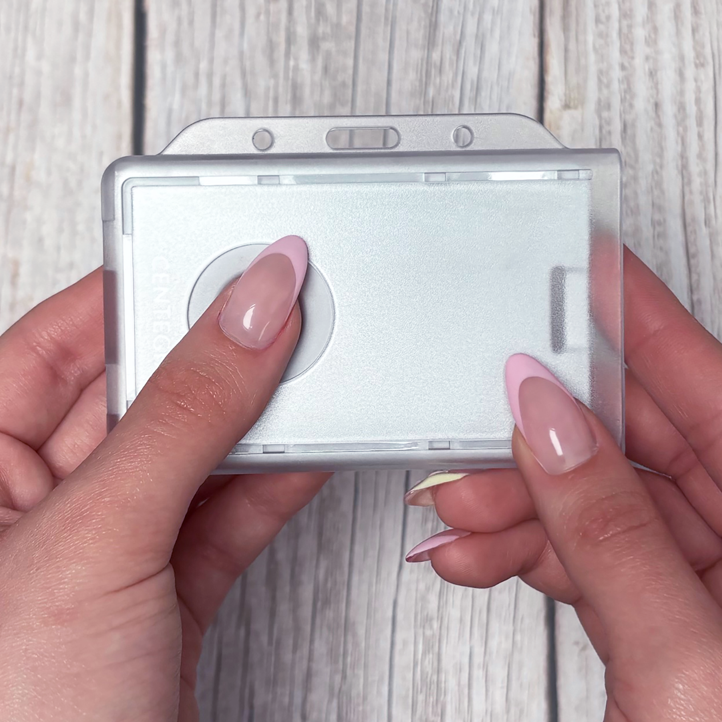 A person with pink and white manicured nails holds a blank, translucent Dual Badge Holder Compatible with Centegix V2 Alarm Card + Standard ID Card above a light wooden surface.