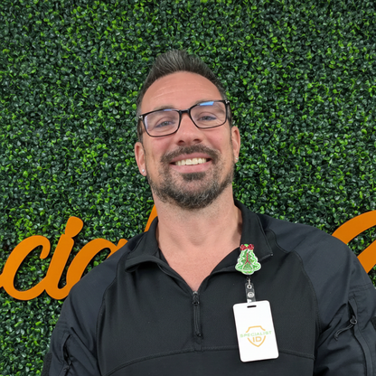 A man in glasses and a black jacket stands before a green leafy wall with orange lettering, smiling at the camera. He has the Holiday Christmas Tree Badge Reel – Cute Festive ID Holder for Nurses, Staff & Professionals clipped to his jacket.
