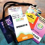 A black lanyard holds several Custom Printed Event Badges with Three Slot Holes— Design It Your Way!—featuring colorful labels for music, art, business, and guest identification on a wooden surface.