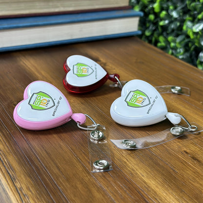 Three Custom Heart Shaped Badge Reels With Rotating Spring Clips - Personalize with Your Logo, in pink, red, and white colors, rest on a wooden surface. Each retractable reel has a slot for attaching a card and displays a logo reading "SPO No." These custom badge reels are perfect to promote brand awareness while adding an element of charm.