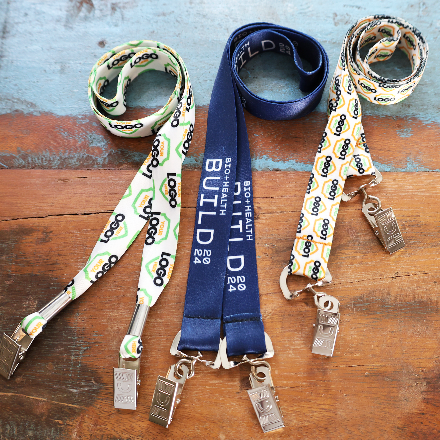Three Custom Double Ended Lanyards with metal clips are shown on wood; two have a "Logo" pattern, and one reads "BioHealth Build 2024," all featuring full color dye sublimation for personalized branding.