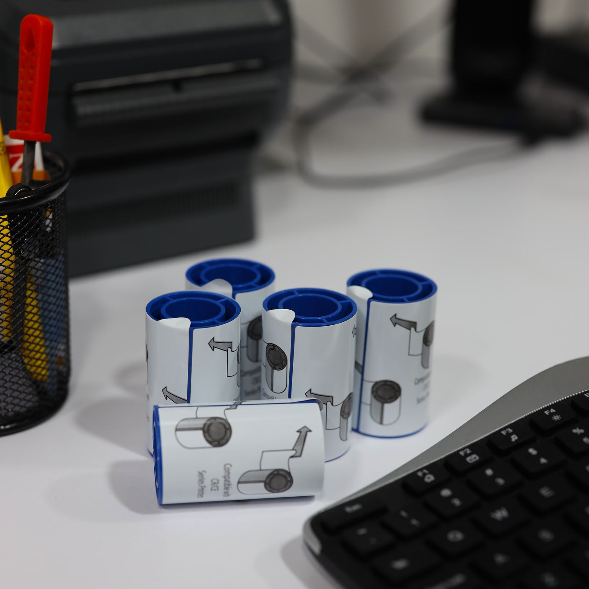 Five Entrust (Datacard) 569946-002 adhesive cleaning sleeves are arranged on a white desk beside a black keyboard and a pen holder, ready for use with your ID card printer—ideal for maintaining printhead protection during frequent printing tasks.