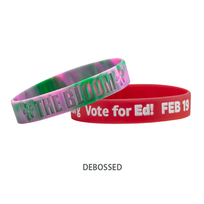 Two debossed, high-quality Customizable Adult 1/2" Silicone Wristbands: one pink and green with "THE BLOOM" text, promoting a cause, and the other red with "My Vote for Ed! FEB 19" text.