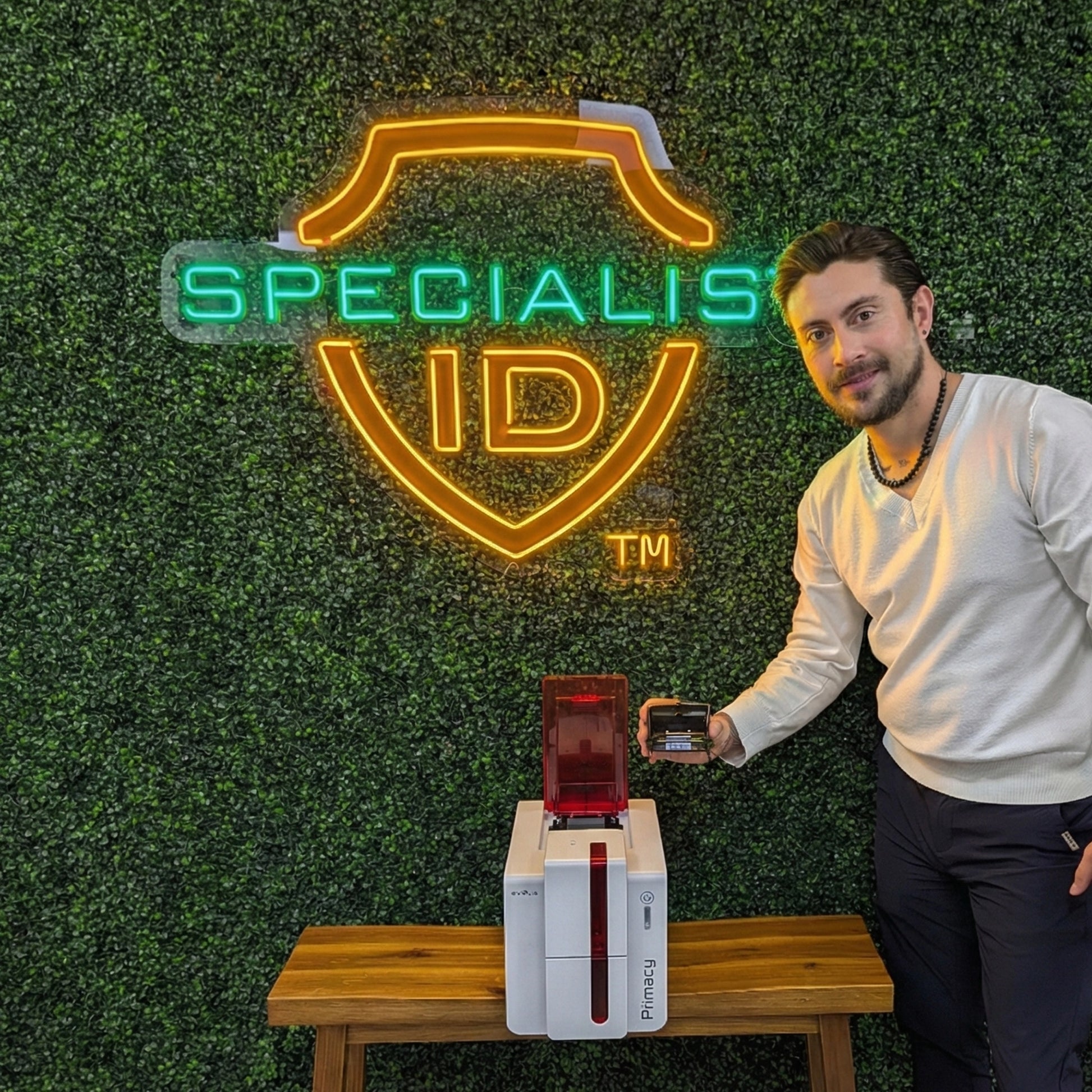 A man stands next to an Evolis S10084 Replacement Printhead for Zenius & Primacy ID Card Printers, holding an ID card, in front of a neon "Specialist ID" sign on a green plant wall.