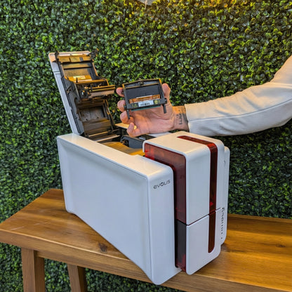 A person holds an Evolis S10084 Replacement Printhead for Zenius & Primacy ID Card Printers above an open printer on a wooden table, with a green plant wall in the background.