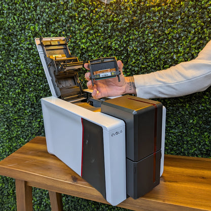 A person installs an Evolis S10381 Replacement Printhead into a Primacy 2 ID Card Printer on a wooden table, with a green leafy wall in the background, emphasizing genuine Evolis parts for reliable performance.