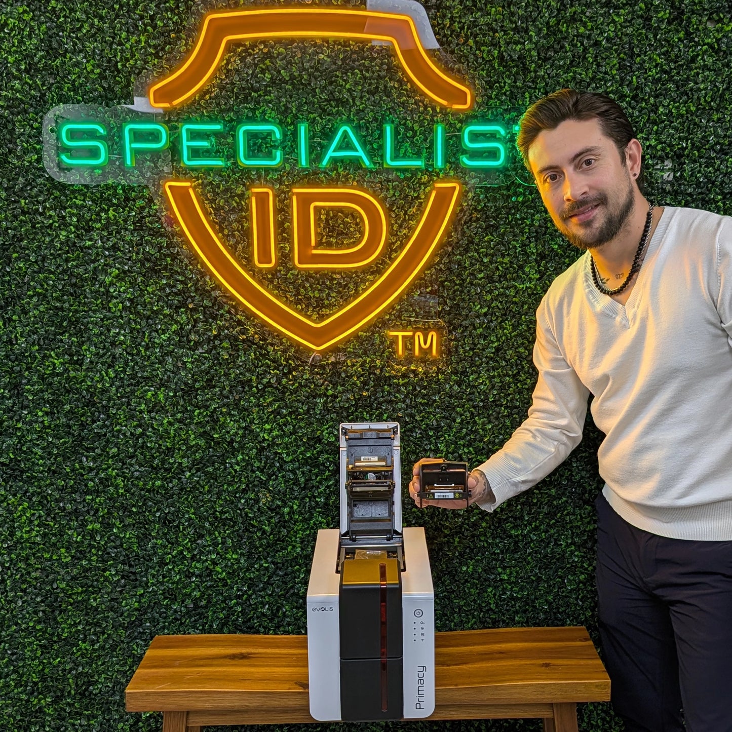 A man stands next to a Primacy 2 printer with an Evolis S10381 Replacement Printhead, holding a card printer ribbon by a table in front of a "SPECIALIS ID" neon sign against a green wall.
