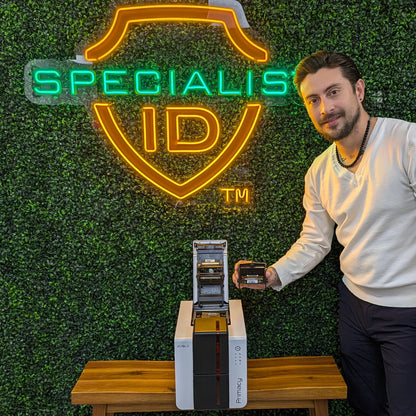 A man stands next to a Primacy 2 printer with an Evolis S10381 Replacement Printhead, holding a card printer ribbon by a table in front of a "SPECIALIS ID" neon sign against a green wall.