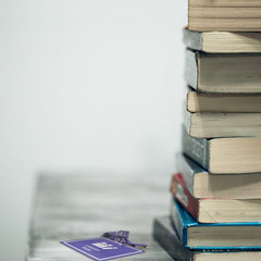 A stack of seven books sits on a table. To the left, Custom Hall Pass Badges - Design Your Own Passes for School, Hospital, Events by Specialist ID with a white logo and text is partially visible. The background is blank and white, highlighting the durable composite material used in both the hall pass badges and some book covers.