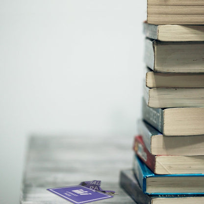 A stack of seven books sits on a table. To the left, Custom Hall Pass Badges - Design Your Own Passes for School, Hospital, Events by Specialist ID with a white logo and text is partially visible. The background is blank and white, highlighting the durable composite material used in both the hall pass badges and some book covers.