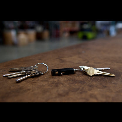 Two sets of keys lie on a wooden surface. One set has multiple keys on a Key-Bak #1101 Quick Release Pull-Apart Key Ring for easy organization, while the other set has three keys and a small black device. A blurred background with indistinct objects is visible.