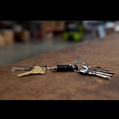 A set of keys lies on a brown surface, featuring a Key-Bak #1101 Quick Release Pull-Apart Key Ring that allows you to easily separate vehicle and house keys. The background is out of focus, displaying an indoor area with indistinct objects.
