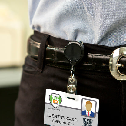 Close-up of a person wearing dark pants and a light shirt, with an ID badge holder attached to a MINI-BAK #67 Black Retractable Reel with 36” Nylon Cord, Vinyl ID Strap and Swivel Clip 0067-005 clipped to their belt. The card displays a photo, the title "Specialist," and a QR code.
