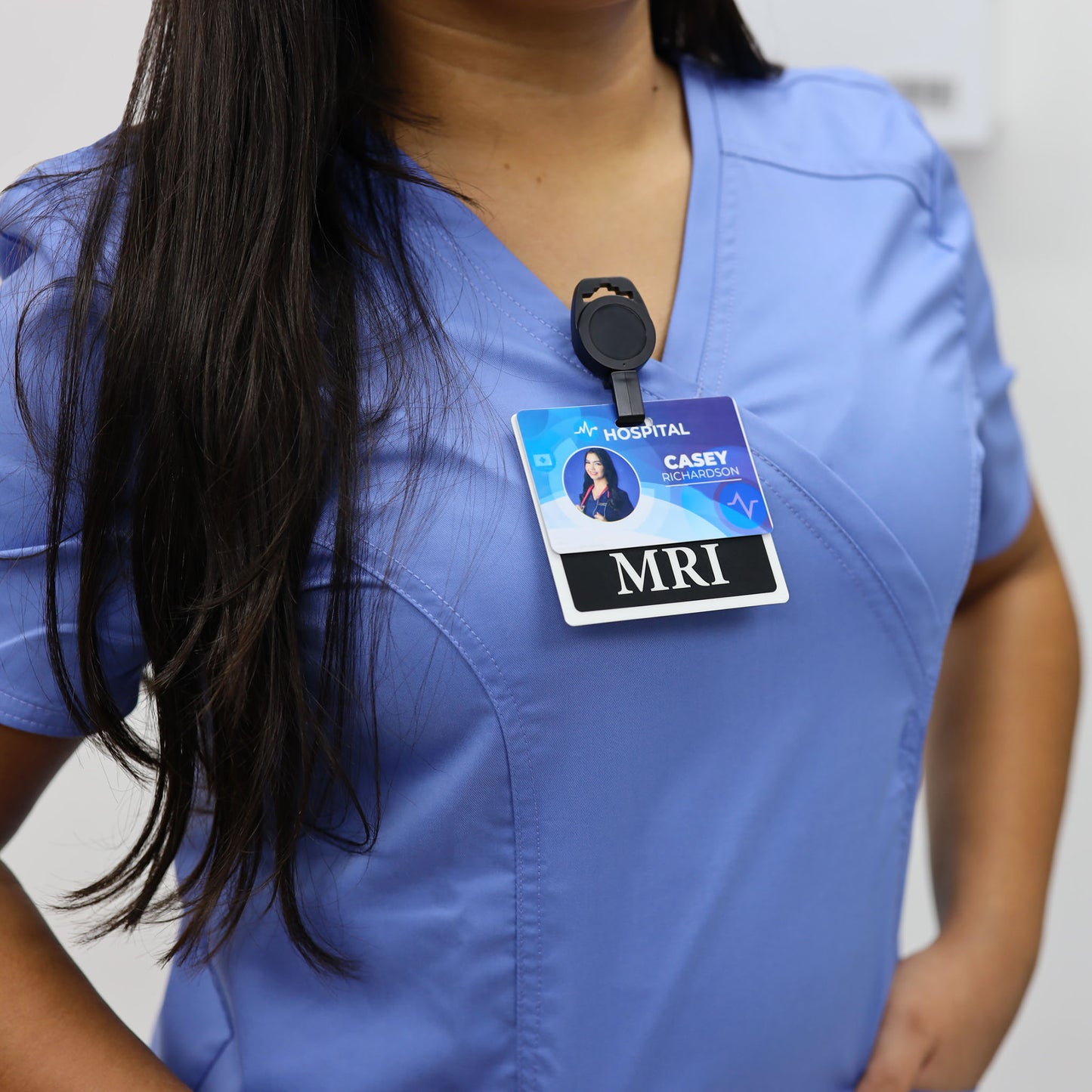 A close-up of a person wearing light blue medical scrubs with a black retractable no twist ID holder clip for imaging room techs & nurses clipped to their chest holding a hospital ID card that reads MRI and CASEY.