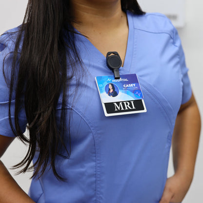 A close-up of a person wearing light blue medical scrubs with a black retractable no twist ID holder clip for imaging room techs & nurses clipped to their chest holding a hospital ID card that reads MRI and CASEY.