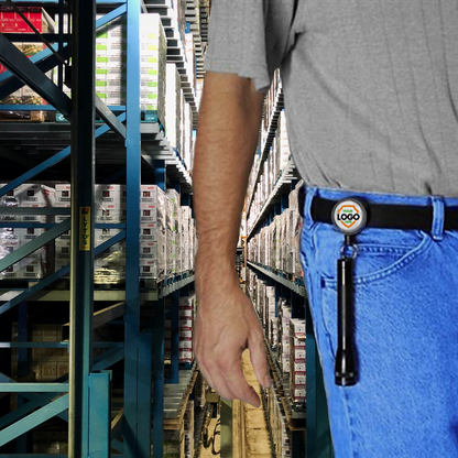 A person in a gray shirt and blue jeans stands in a warehouse aisle, next to tall shelving units stacked with boxes. The person has a flashlight clipped to their belt and is using Customizable Key-Bak Mid Size Key Ring Badge Reel with Belt Clip (#6) - Add Your Logo to promote brand awareness.