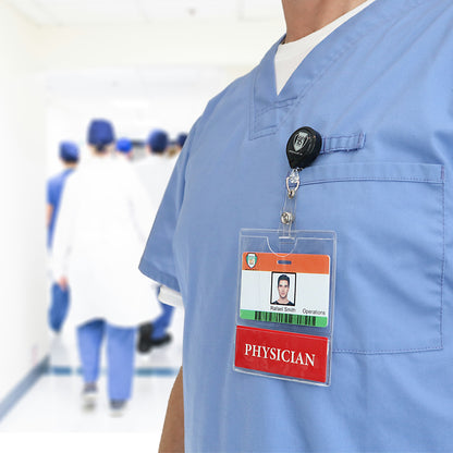 A person wearing blue scrubs and a PHYSICIAN BadgeBottom Badge Holder & PHYSICIAN Badge Buddy IN ONE!! - Horizontal ID Badge Sleeve with Bottom Role Tag for Nurses with a visible ID badge labeled "Physician" stands in a hospital hallway, with several other people in medical attire walking in the background.