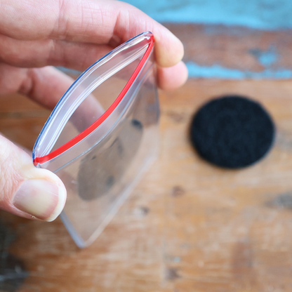 A hand holds an open Heavy Duty Resealable Badge Holder with Adhesive Hook & Loop Back (PPP-1) on a wooden surface. A round black object is in the background.