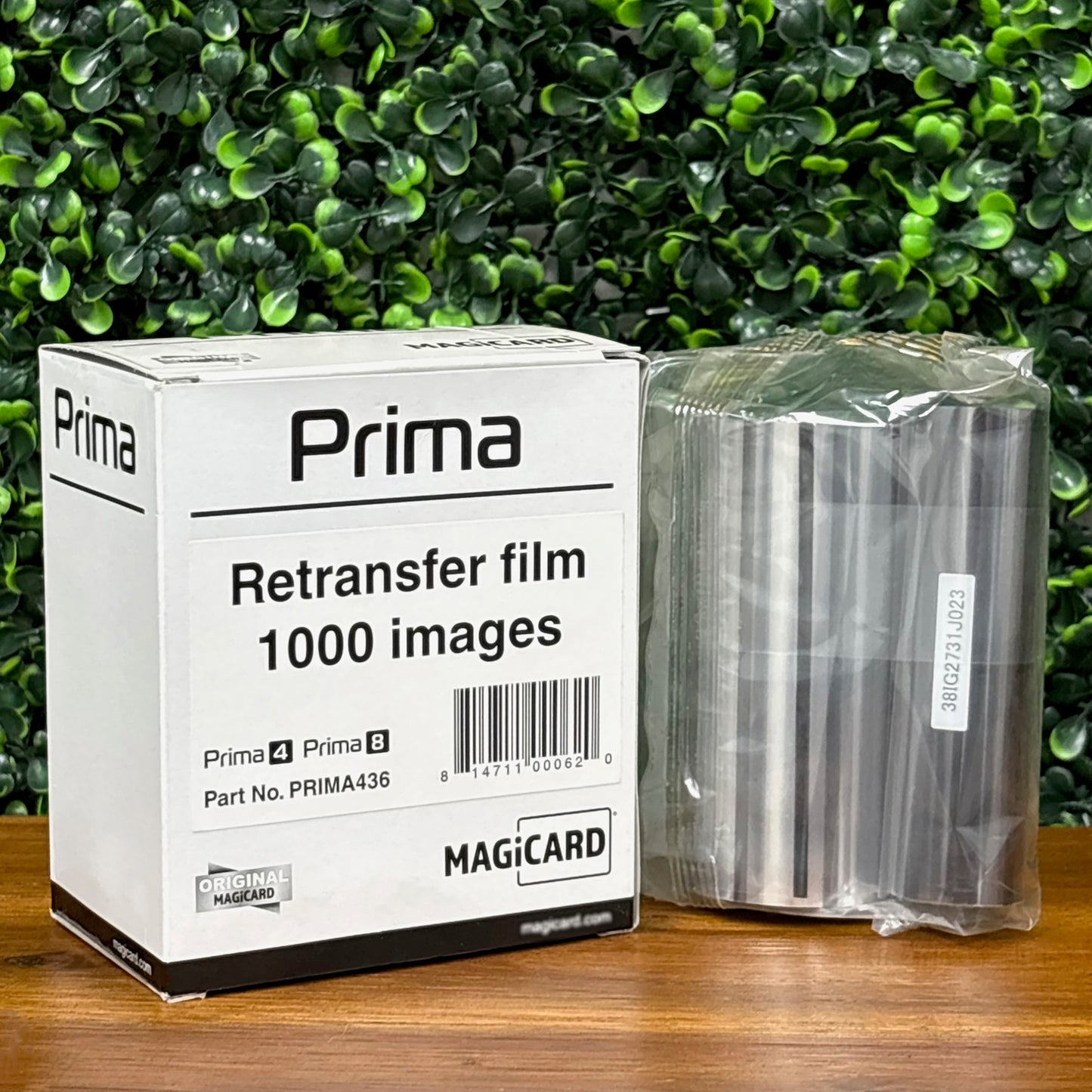 A box labeled "Magicard PRIMA436 Retransfer Film for the Magicard Prima 4 and Prima 8, 1,000 Prints" and a sealed roll of retransfer film rest on a wooden surface with a green leafy background.