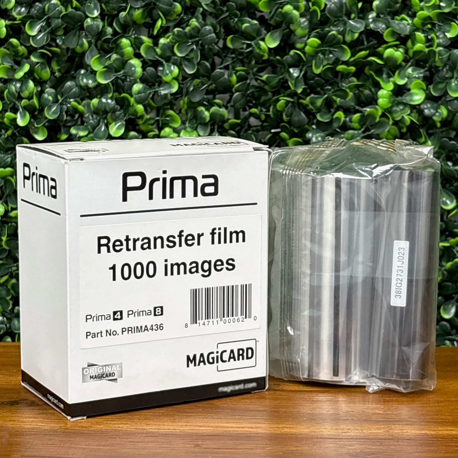 A box labeled "Magicard PRIMA436 Retransfer Film for the Magicard Prima 4 and Prima 8, 1,000 Prints" and a sealed roll of retransfer film rest on a wooden surface with a green leafy background.