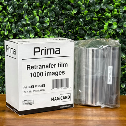 A box labeled "Magicard PRIMA436 Retransfer Film for the Magicard Prima 4 and Prima 8, 1,000 Prints" and a sealed roll of retransfer film rest on a wooden surface with a green leafy background.
