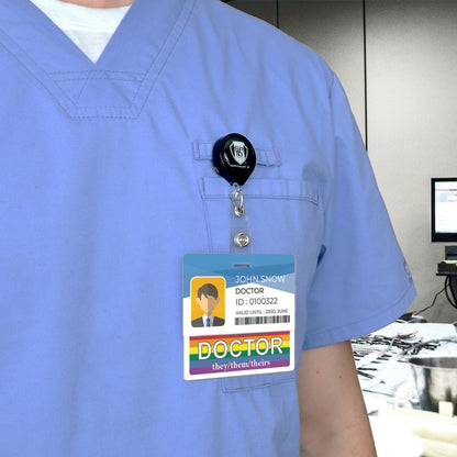 A person wearing a blue scrub top with a DOCTOR Pronouns Badge Buddy Horizontal with Rainbow Border displaying the name "John Snow, Doctor," a photo, ID number, and pronouns "they/them/theirs" in a medical setting with monitors in the background. A subtle rainbow pride pin adorns their chest pocket.