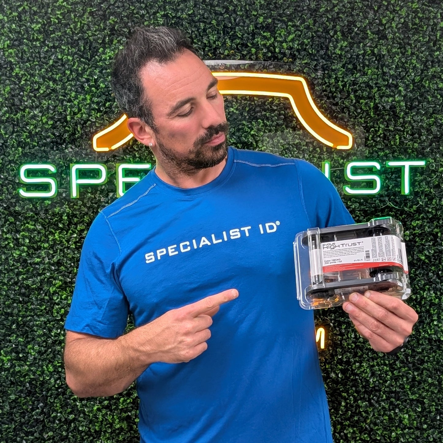 A man in a blue "Specialist ID" shirt points to a clear case holding the Evolis R5F002AAA YMCKO HighTrust® Color Ribbon (200 Prints), used in Zenius and Primacy printers, standing before a green wall marked "SPECIALIST.
