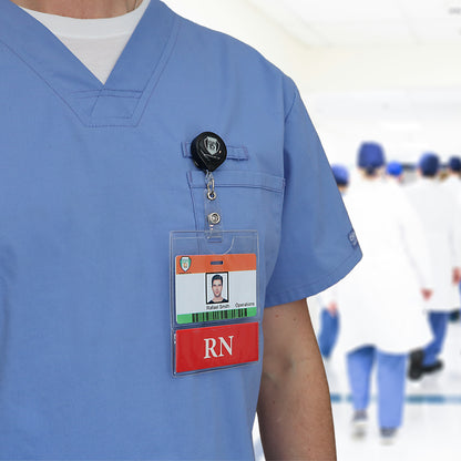 A person in blue scrubs is wearing an RN BadgeBottom Badge Holder & RN Badge Buddy IN ONE!! - Horizontal ID Badge Sleeve with Bottom Role Tag for Registered Nurses. The badge has a photo, text, and an "RN" designation. The blurry background shows others in similar attire walking in a hallway, each displaying their own medical titles prominently.
